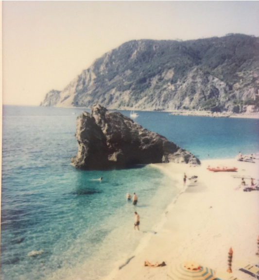 Sun-drenched beach in Cinque Terre, Italy, with turquoise waters, swimmers, striped umbrellas, and dramatic coastal cliffs in the background.