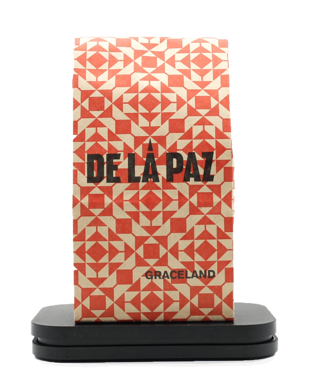A bag of DE LA PAZ coffee by Graceland, featuring a geometric patterned design in orange and cream, displayed on a black stand against a white background.