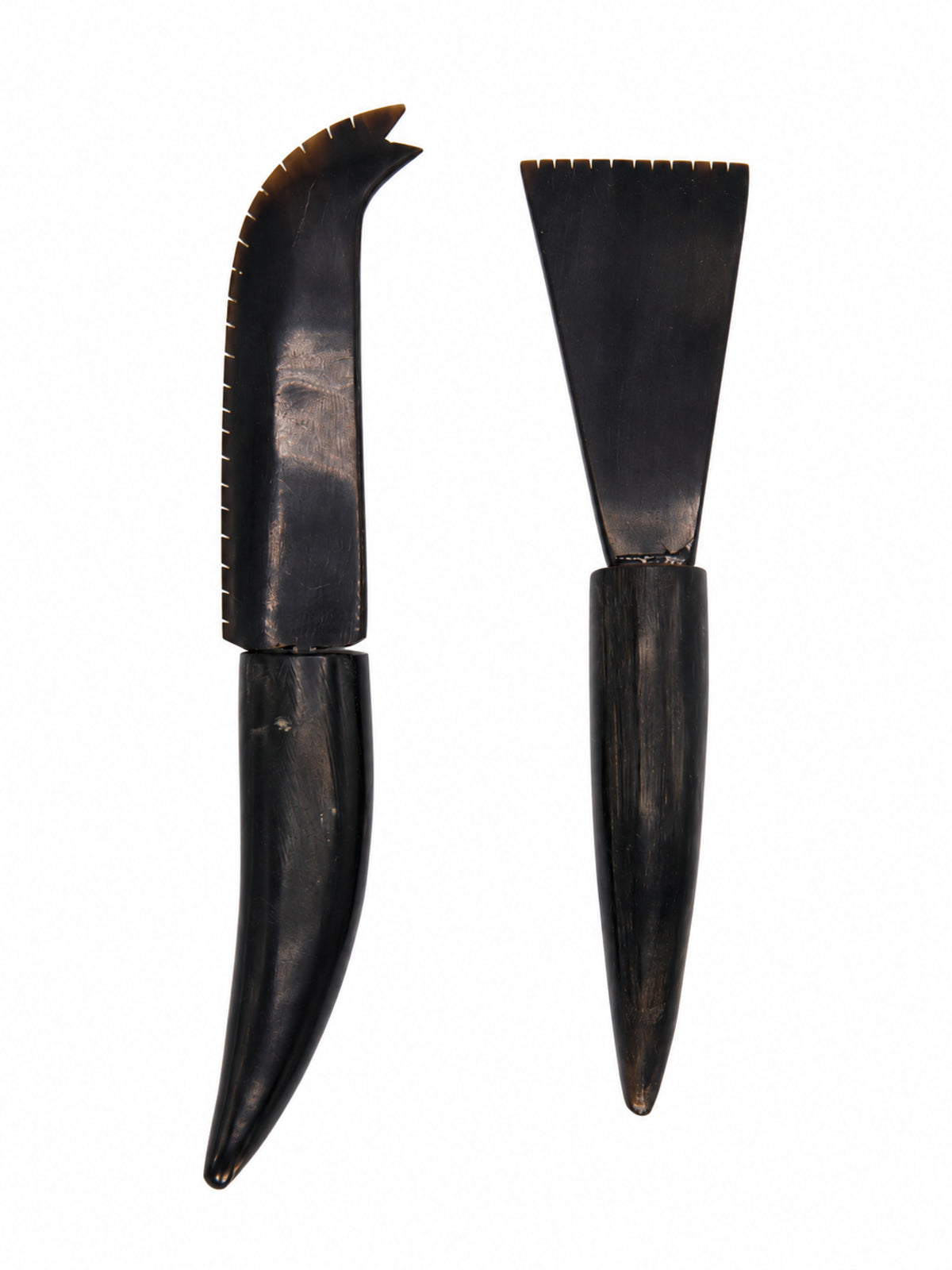 Two dark carved wooden tools with serrated edges, shown upright on a plain white background. One has a curved, hook-like blade, and the other has a wide, flat triangular blade.