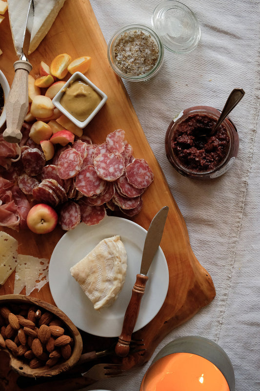Overhead view of an antipasti board on a wooden surface — sliced salumi, soft white cheese on a small plate, stone fruit, whole almonds in a bowl, whole grain mustard, olive spread, and a lit candle on white linen. Styled for summer entertaining.