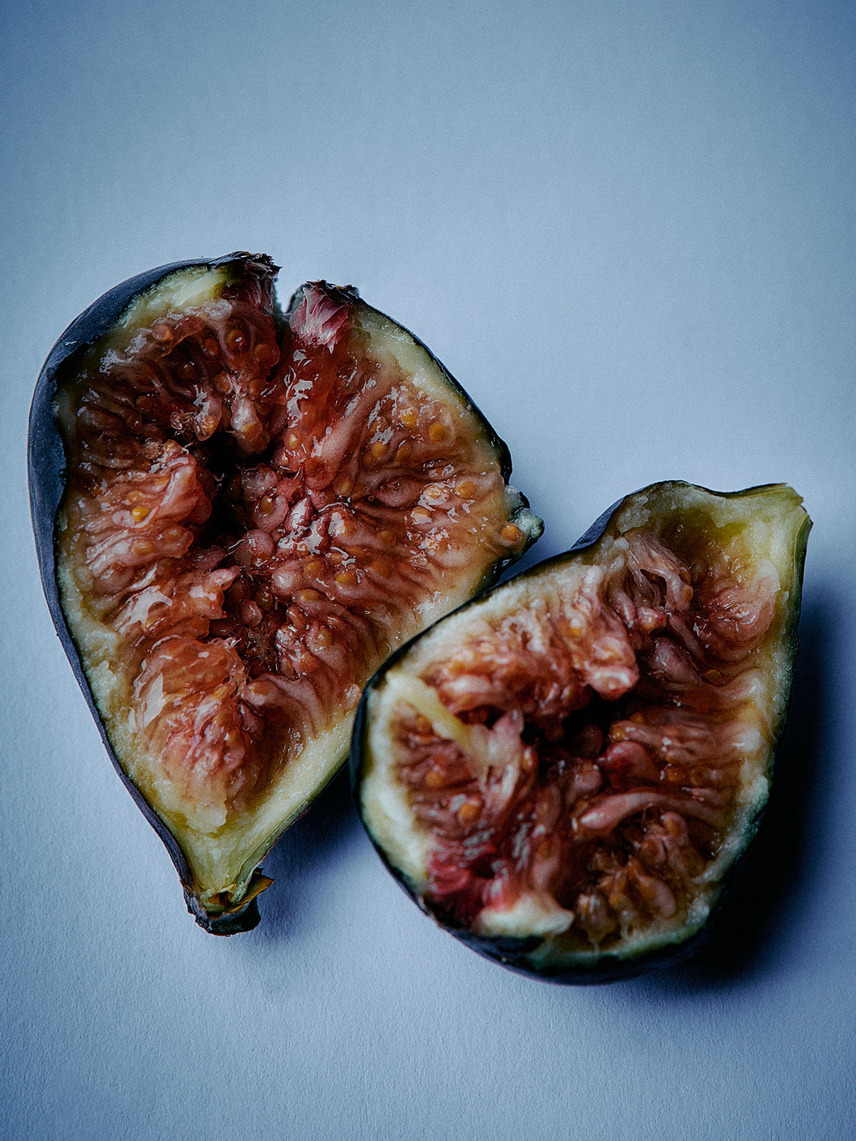Two halves of a fig fruit with pinkish-red flesh and dark green to black outer skin, photographed on a cool blue-gray background.