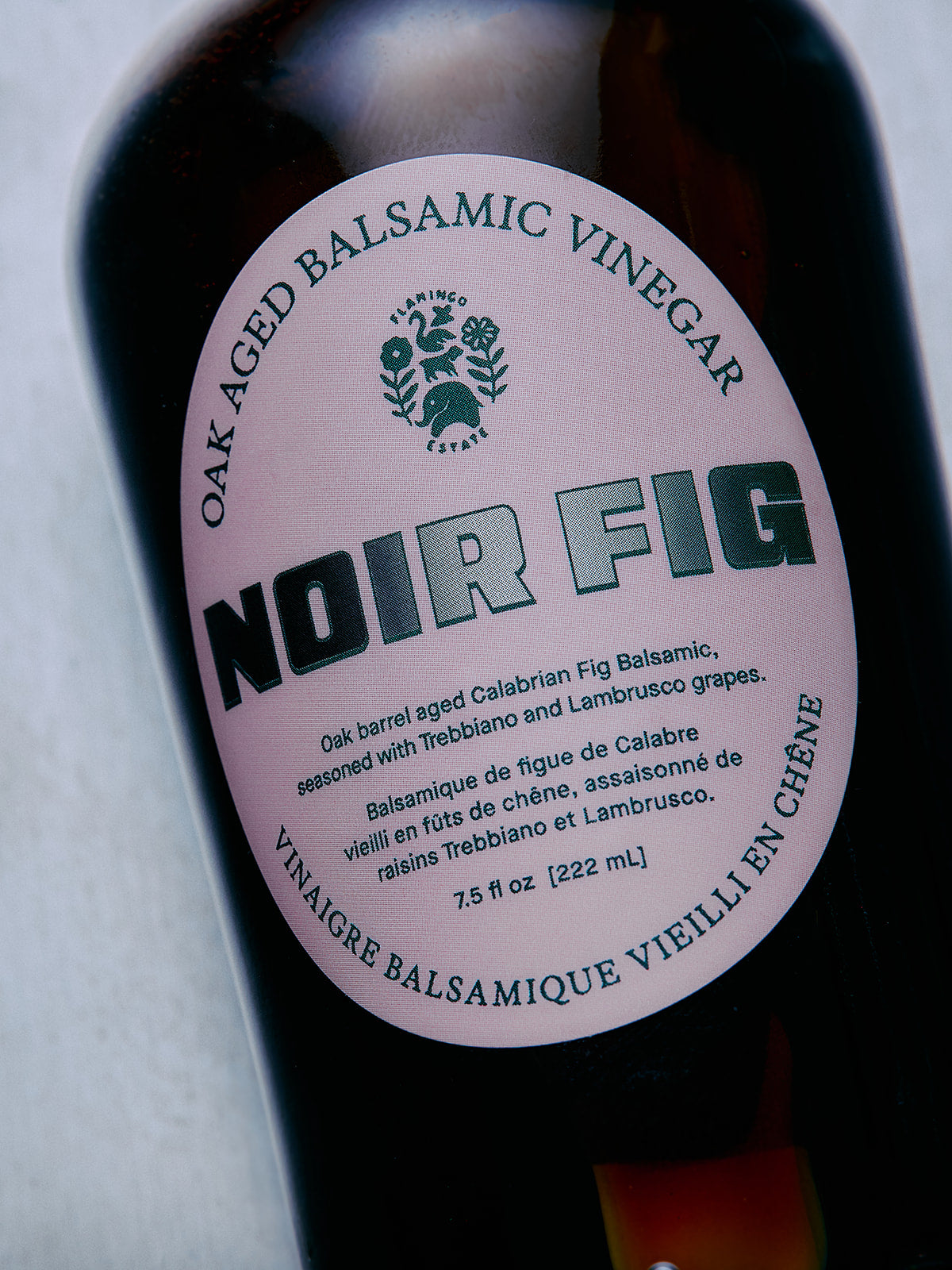 A close-up of a dark bottle of Noir Fig Oak Aged Balsamic Vinegar, showing a round white label with green text and a small crest logo.