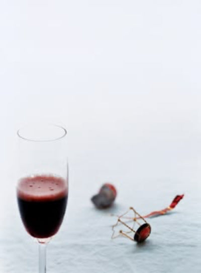 A clear champagne flute filled with deep red wine, photographed on a pale blue surface against a light gray background. A few small red and dark decorative objects lie nearby.