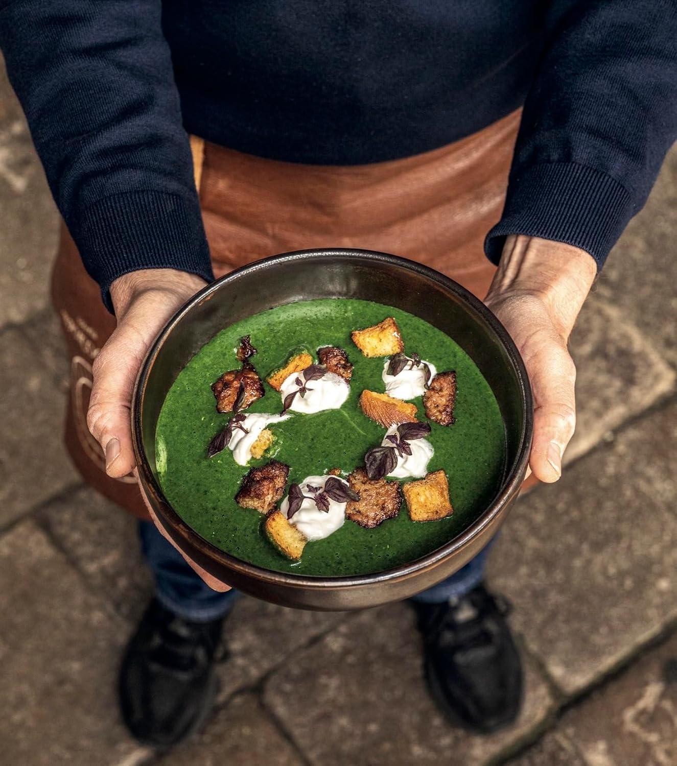 A person in a dark navy sweater and brown apron holds a dark metal bowl filled with a vibrant green soup or sauce, garnished with white cream, chocolate drizzle, and crumbled browned pastry pieces, set against a stone pavement background.