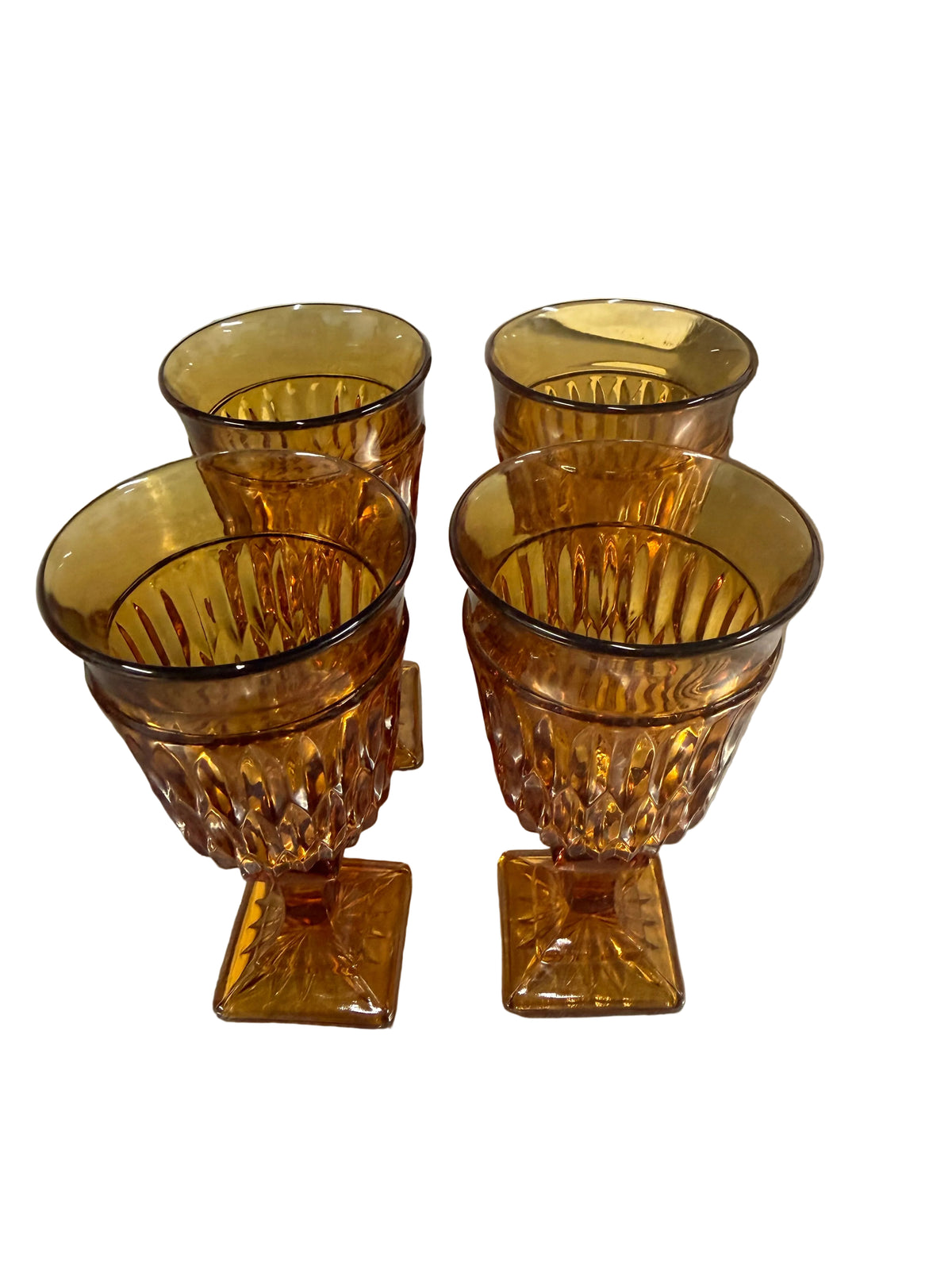 Four amber-colored cut crystal goblets with square bases arranged on a stainless steel countertop, with a glass jar and cardboard boxes in the background.