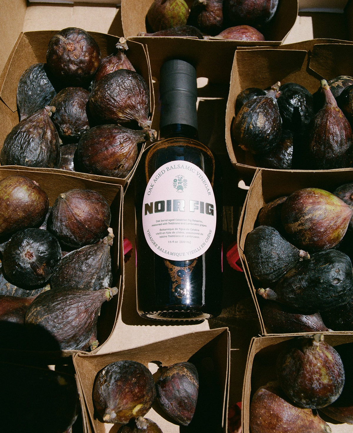 A bottle of Noir Fig wine surrounded by open cardboard boxes filled with dark figs, with the bottle label reading 'NOIR FIG'.