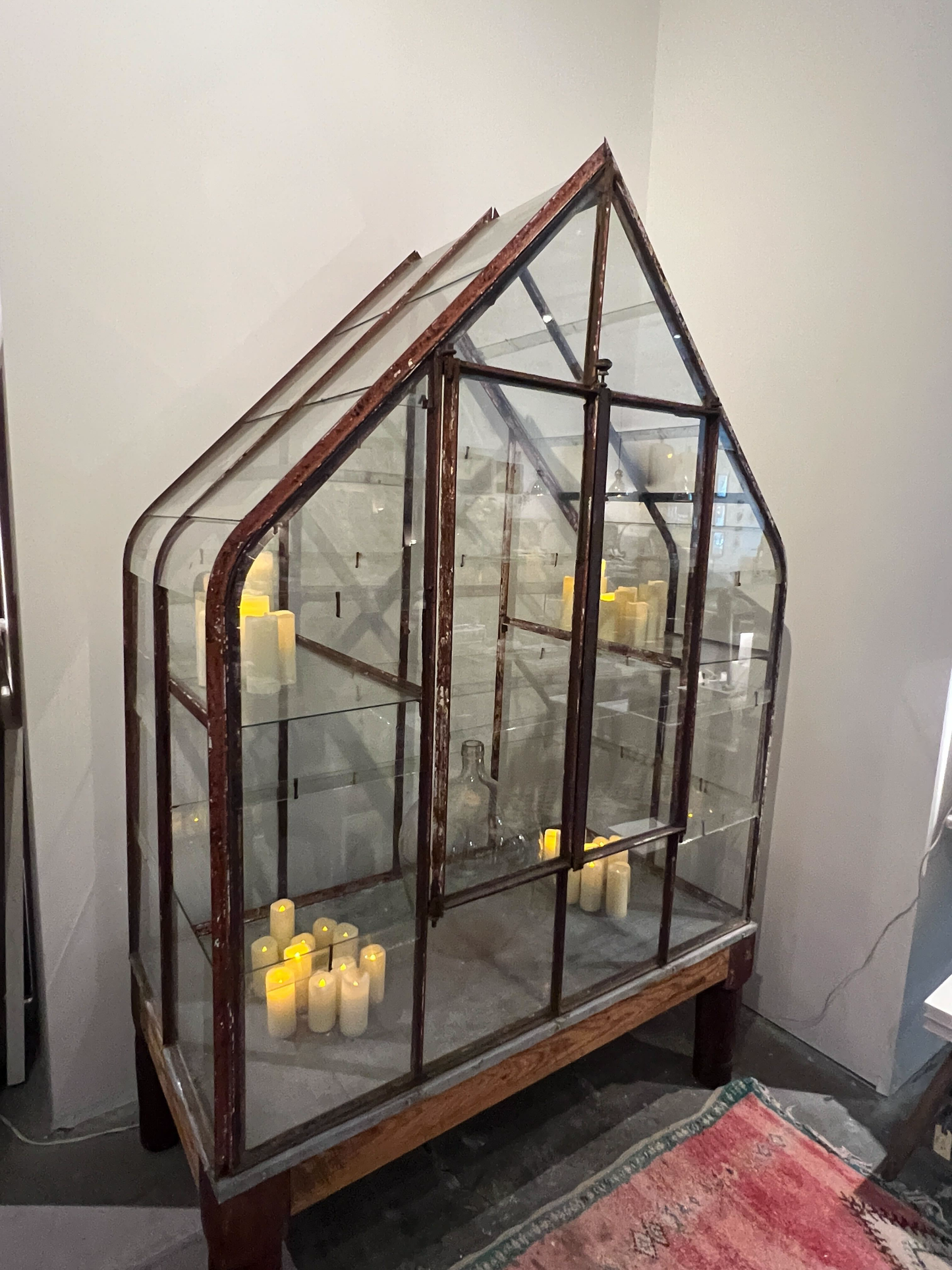 A rustic house-shaped glass display case with a weathered dark brown metal frame and shelves, filled with lit candles, set against a plain light gray wall.