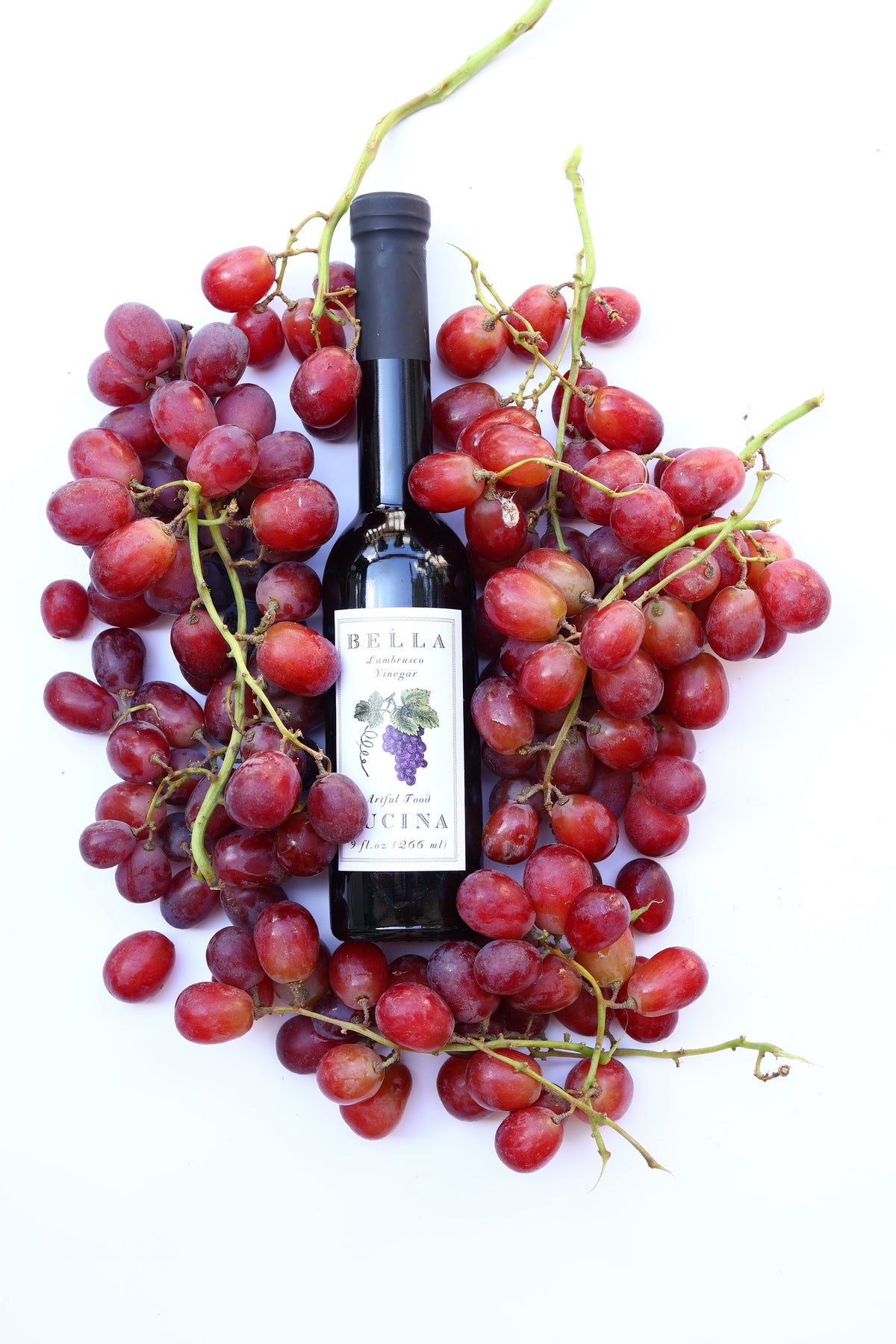 A bottle of Bella Valtellina DOCG white wine with a black cap and a white label, styled on a white background and surrounded by fresh red grapes on the vine.