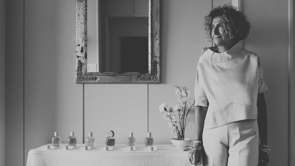 A black and white editorial photograph of a woman standing next to a draped table with perfume bottles and a mirror on a paneled wall.