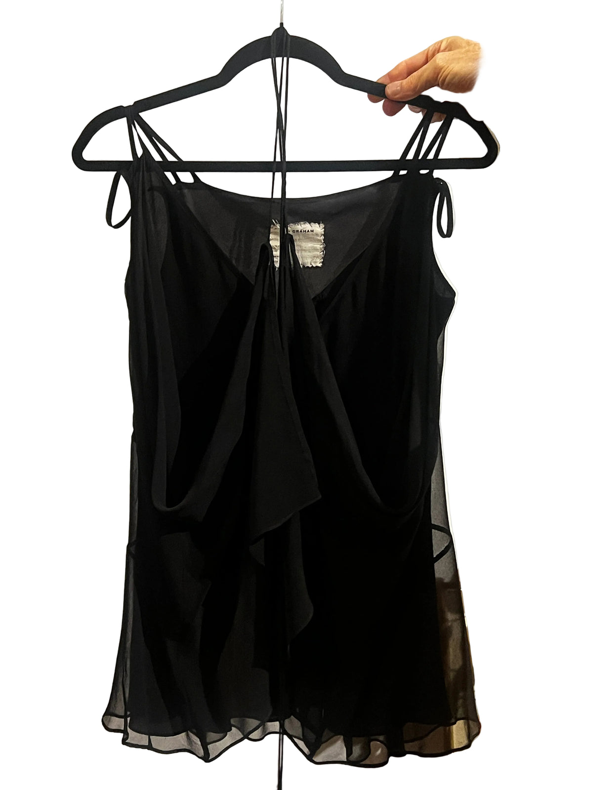 A black sheer sleeveless top with thin shoulder ties and a draped front, hanging on a black hanger held by a person's hand in an indoor setting.