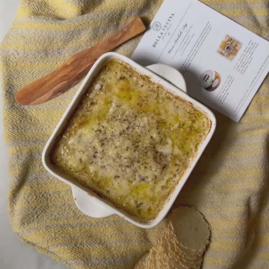 Bella Cucina baked artichoke dip made with Artichoke Lemon Pesto, golden and bubbling in a baking dish with recipe card and wooden spreader. 