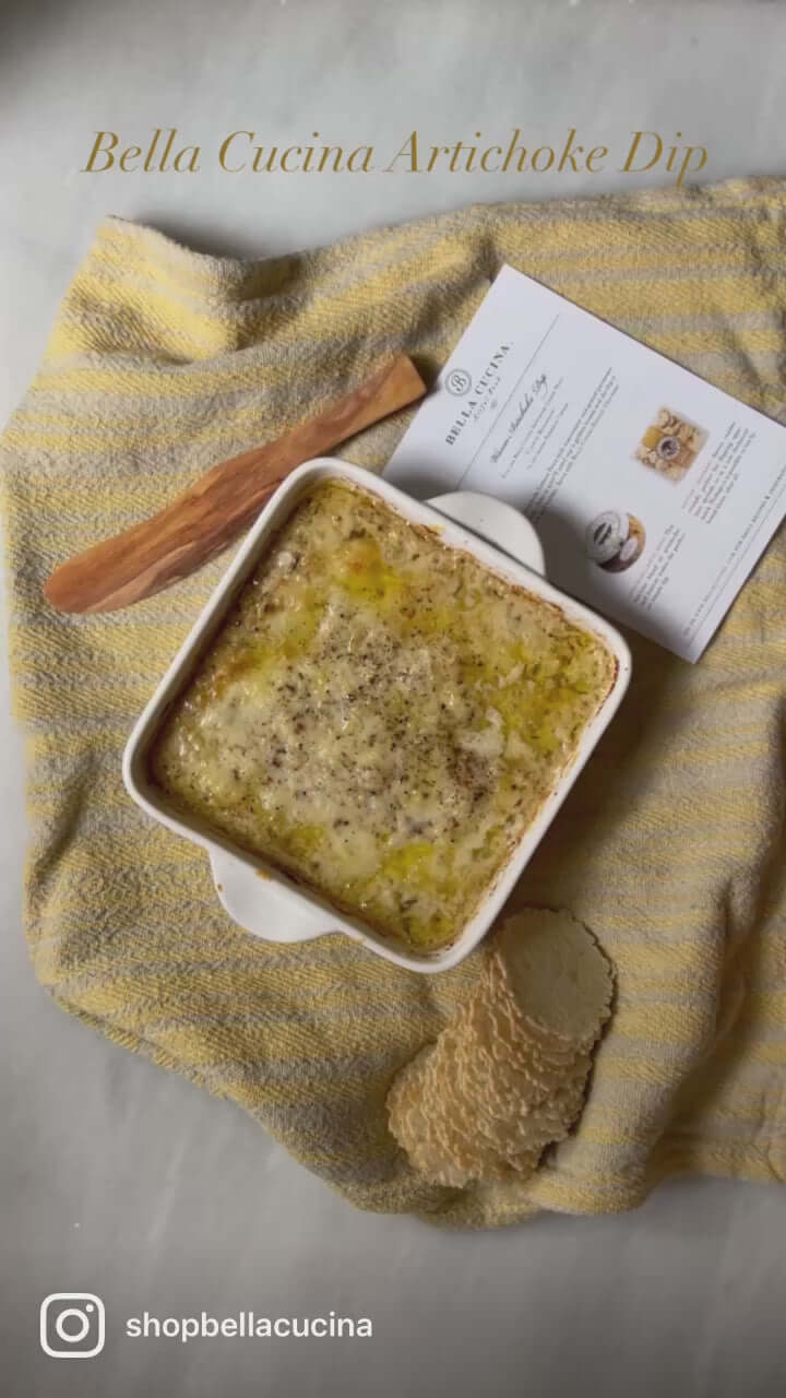 Bella Cucina baked artichoke dip made with Artichoke Lemon Pesto, golden and bubbling in a baking dish with recipe card and wooden spreader. 