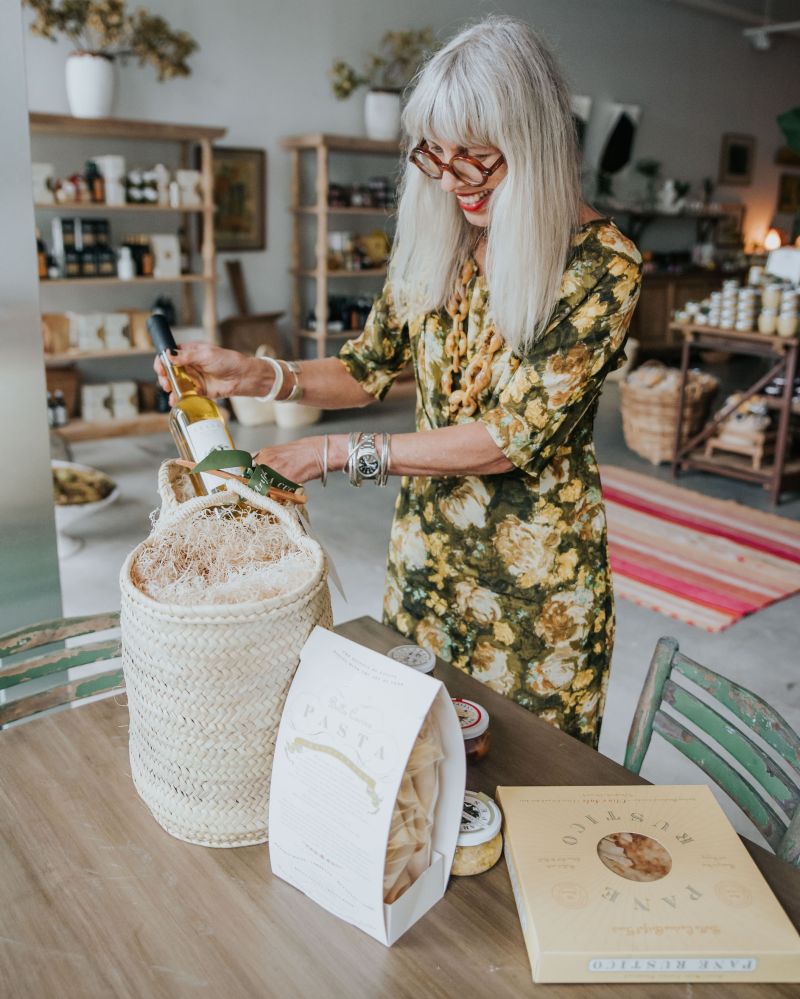 Stylish woman with silver hair, wearing a floral dress, unpacks a gourmet Farmer’s Market Basket at Bella Cucina, featuring artisanal pasta, olive oil, and gourmet treats in a woven basket.