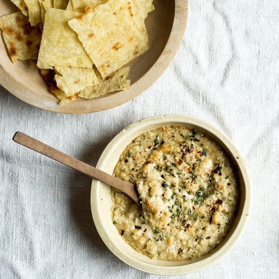 Rustic bowl of creamy Artichoke Lemon Pesto dip, garnished with herbs and red pepper flakes, served with crispy Tuscan crackers in a wooden bowl on a linen tablecloth.