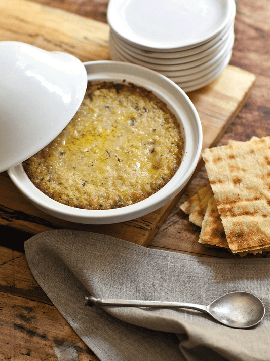 Baked Artichoke Lemon Pesto dip in a white ceramic dish, served with crispy Tuscan crackers on a rustic wooden table, accompanied by a linen napkin and antique silver spoon.