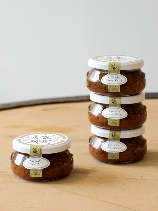 Stacked jars of Bella Cucina Olivada Olive Pesto, a gourmet olive spread made with black and green olives, displayed on a wooden surface.